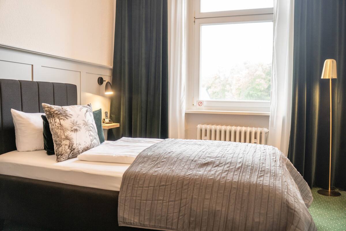 Quarto de hotel com cama, mesa de cabeceira com luminária, janela com cortinas e radiador.