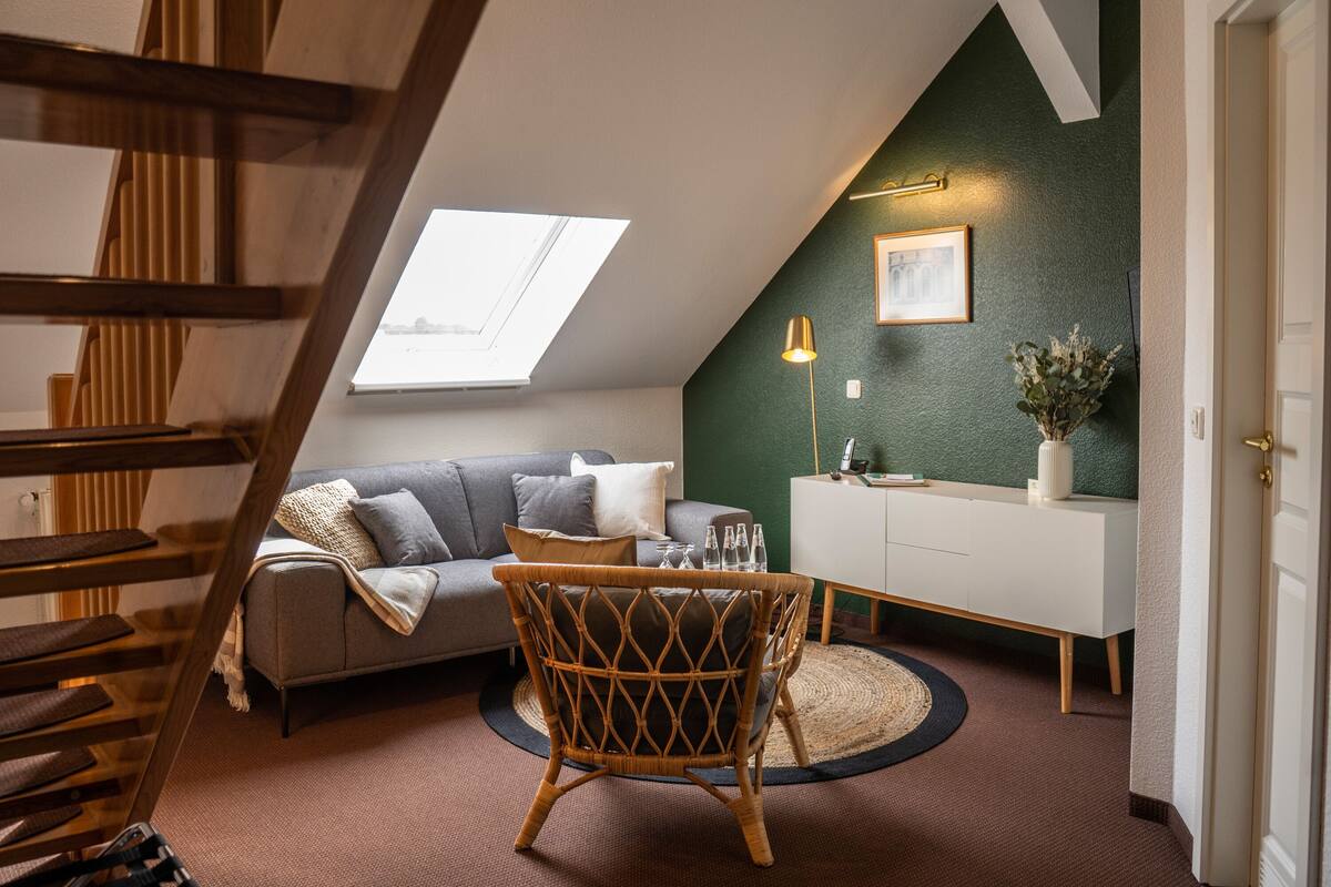 Um quarto em estilo loft com teto inclinado, um sofá cinza, uma poltrona de vime, um armário branco e uma pequena mesa com copos.
