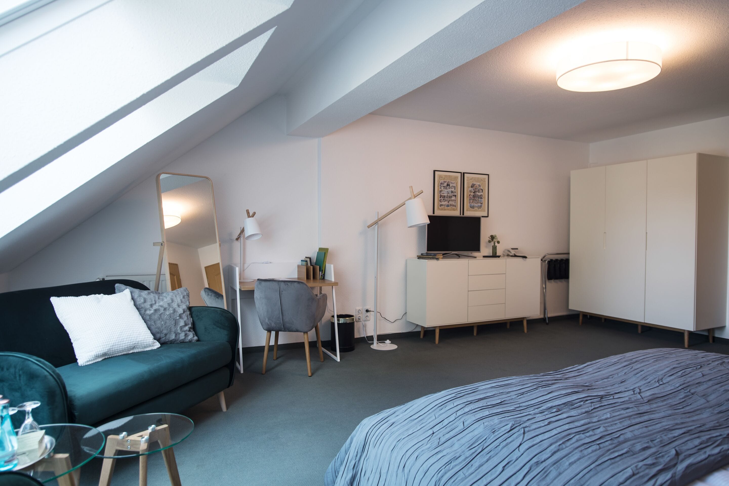 A modern, compact room with a sloped ceiling, a bed, a desk with a chair, a wardrobe, and a large mirror.
