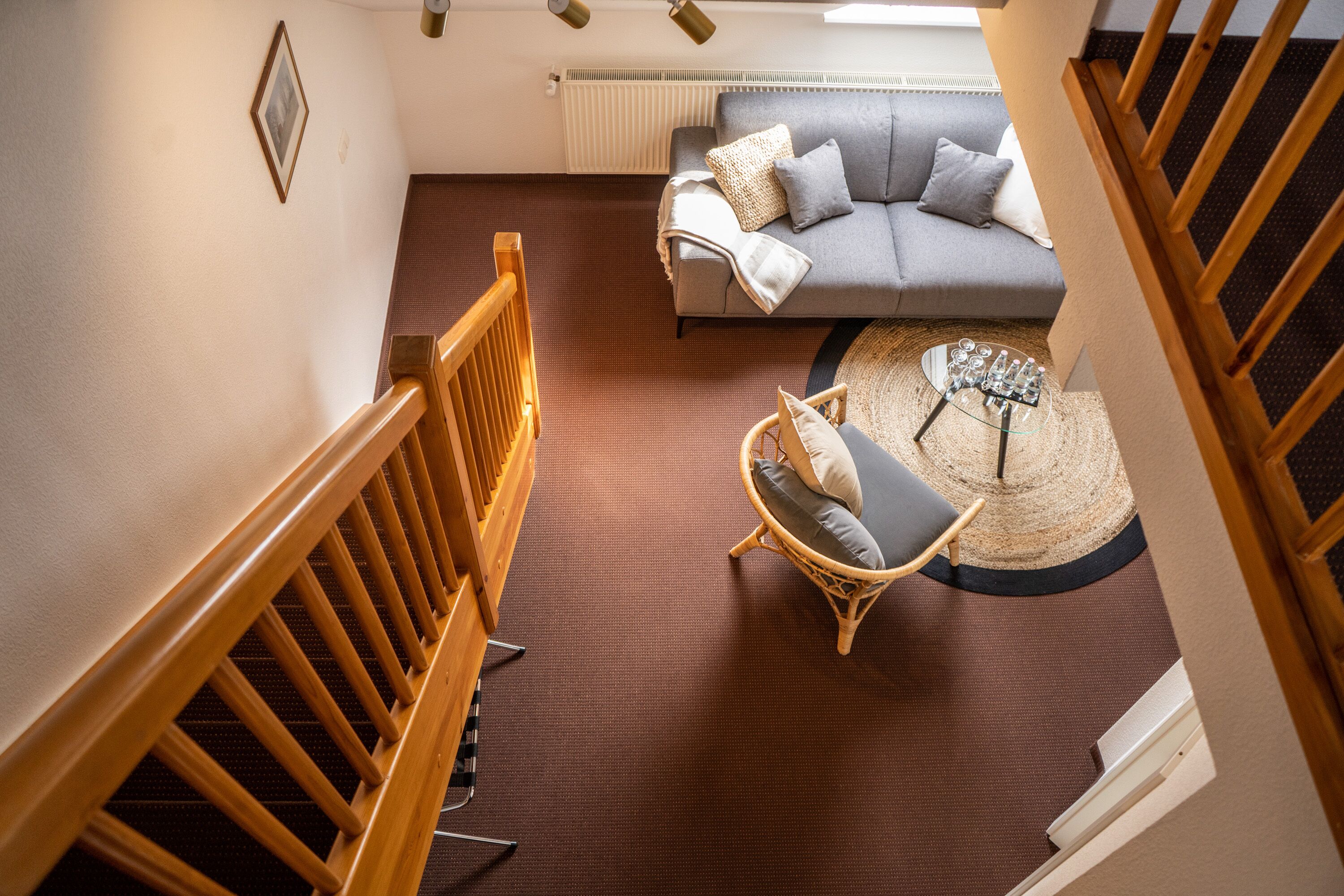 A staircase leading to a living area with a grey sofa, wicker chair, and a round table with glasses.