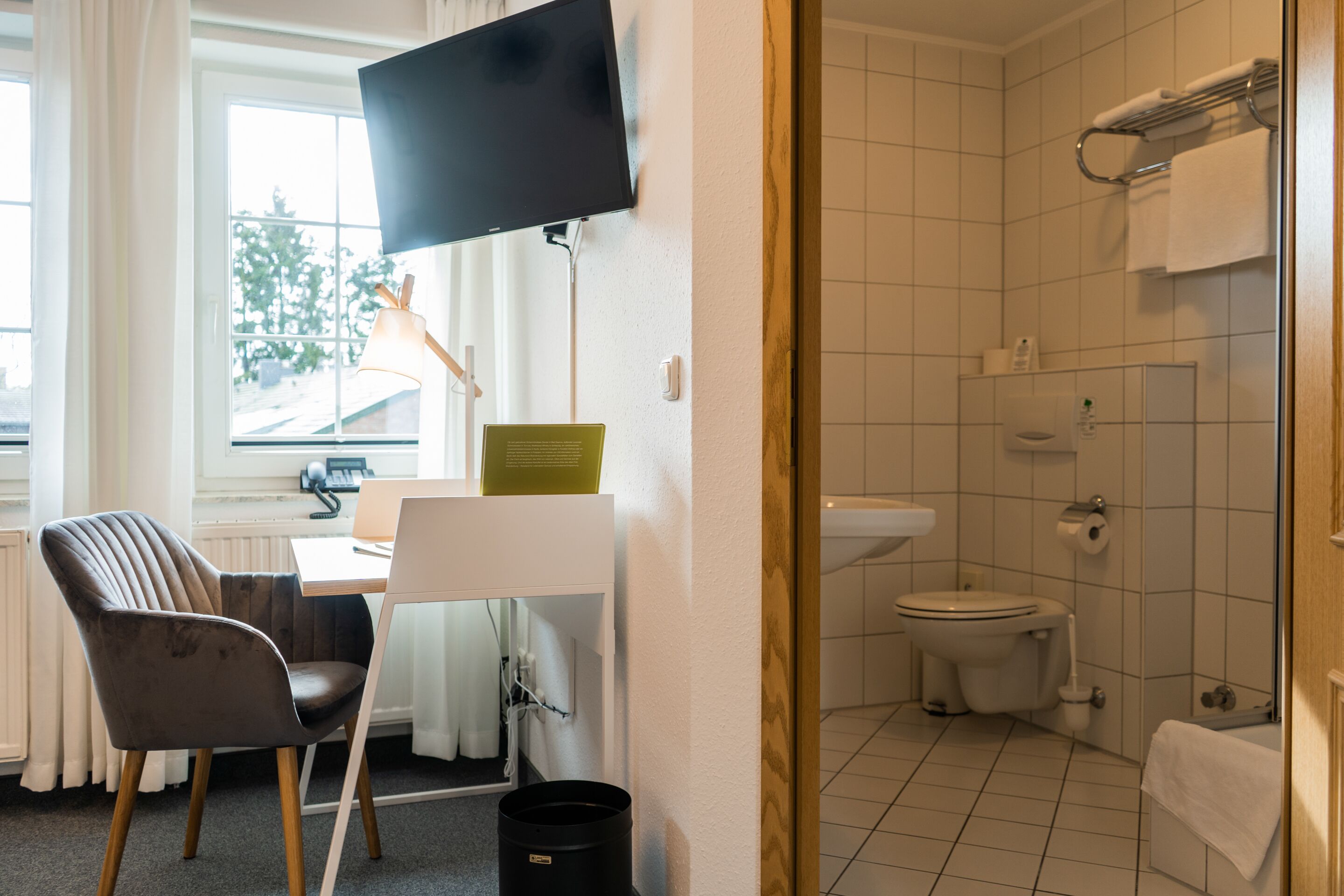 A modern bathroom with a toilet, sink, and shower. There is a chair and a desk with a computer and a lamp. A television is mounted on the wall.