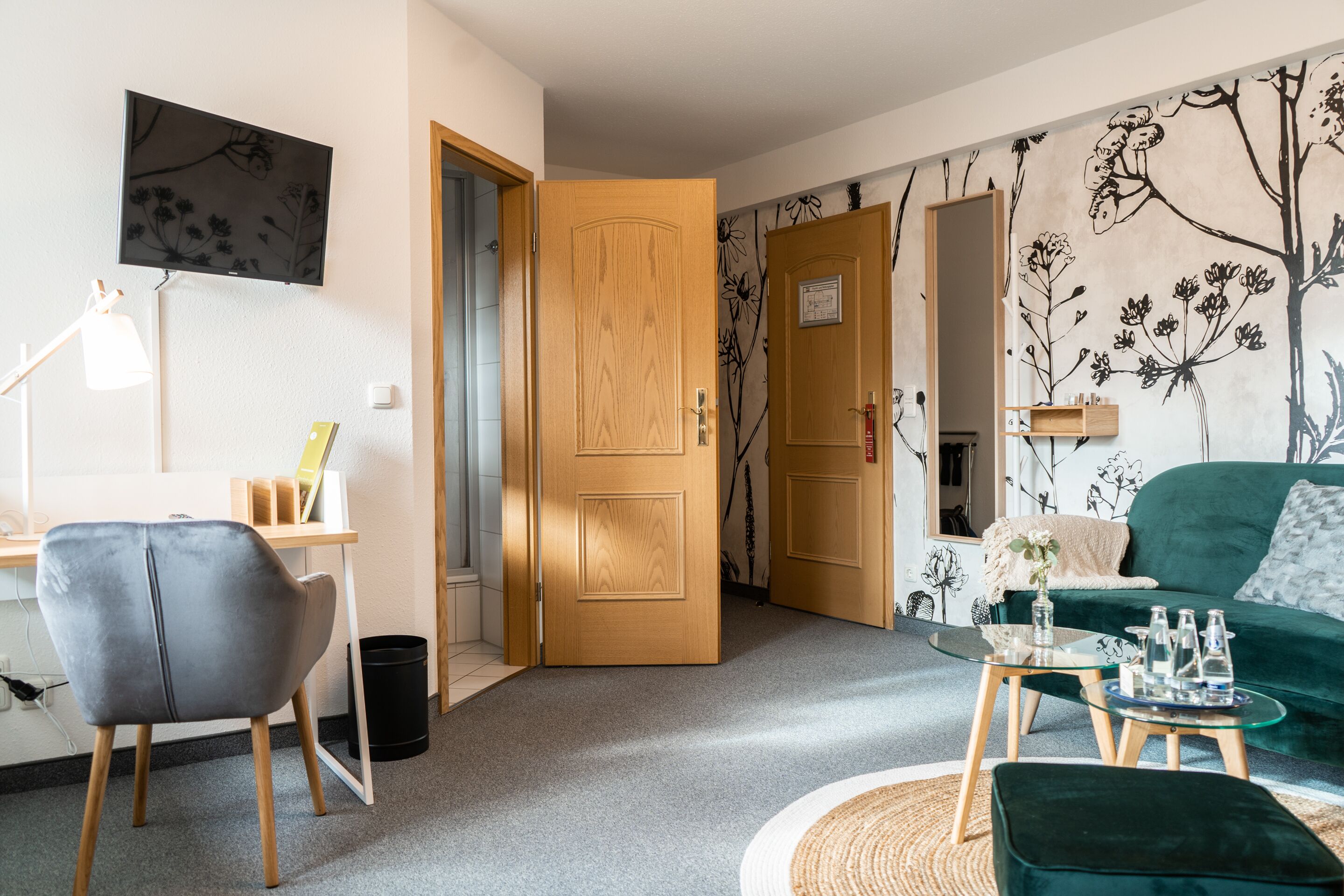 Un salon avec un canapé vert, un fauteuil gris, une porte en bois et un mur recouvert d’un papier peint à motifs floraux.