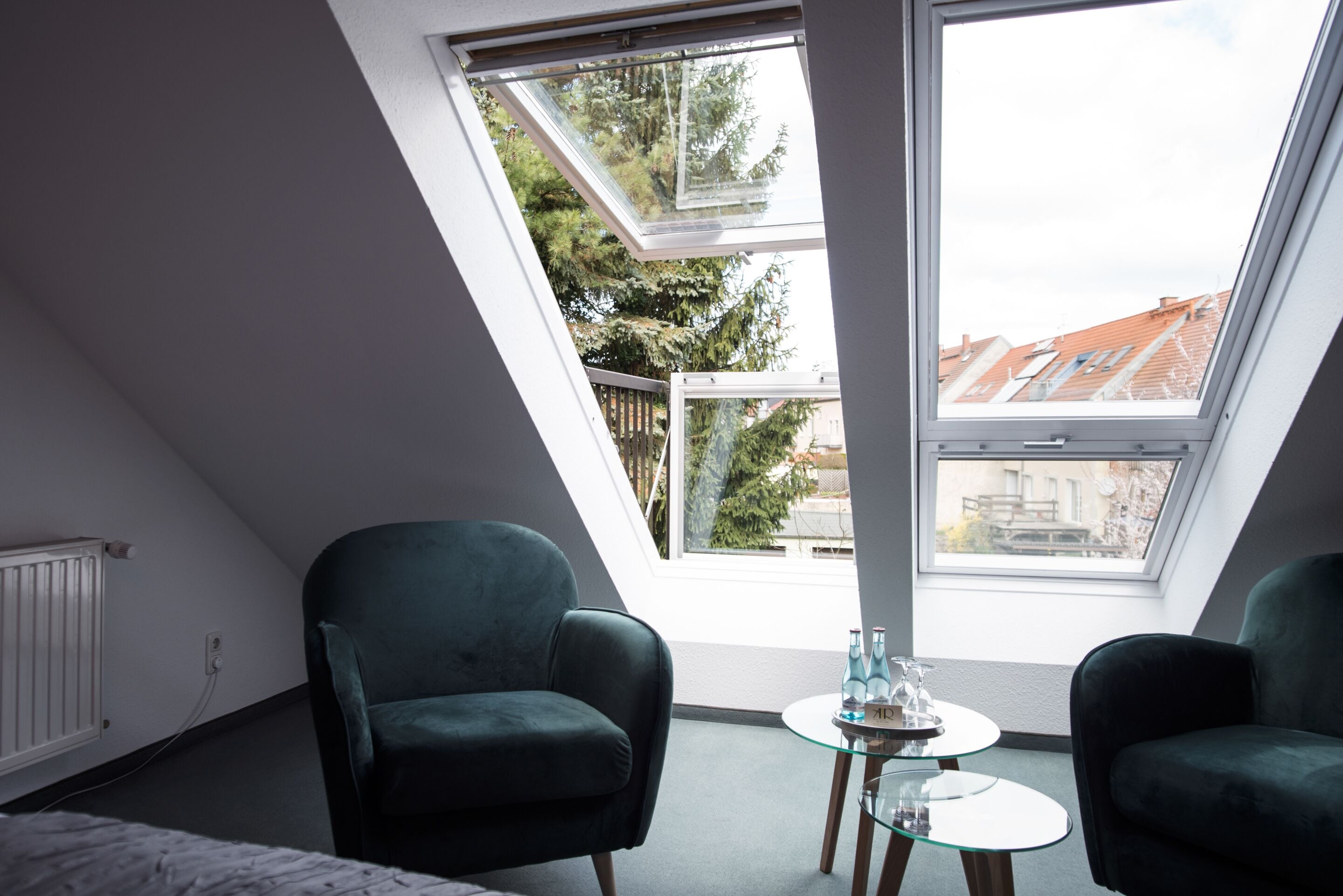A room with a sloped ceiling, two green armchairs, a small glass table with bottles, and a view of trees and buildings through a skylight.