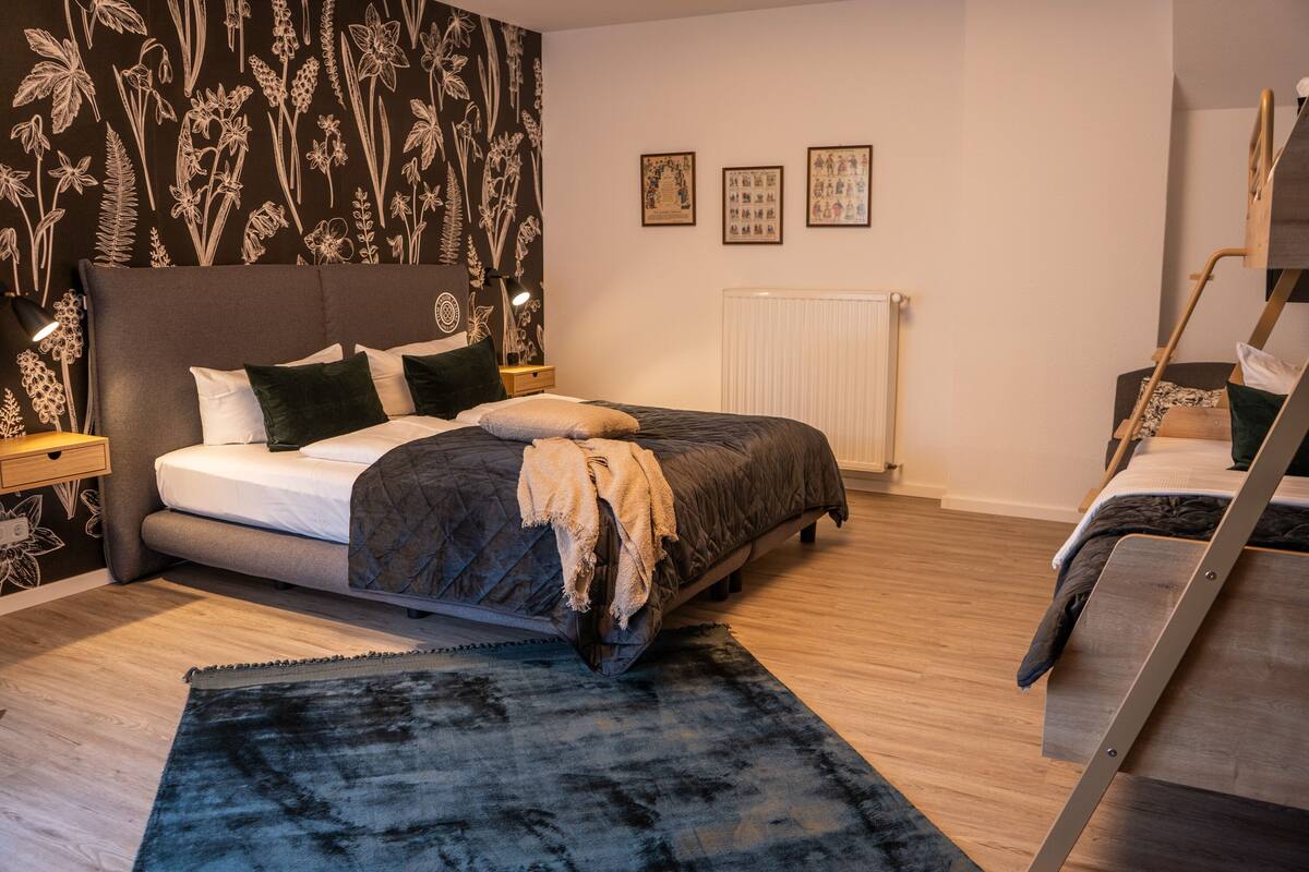 Quarto com cama, mesa de cabeceira, radiador, beliche e parede com papel de parede floral.