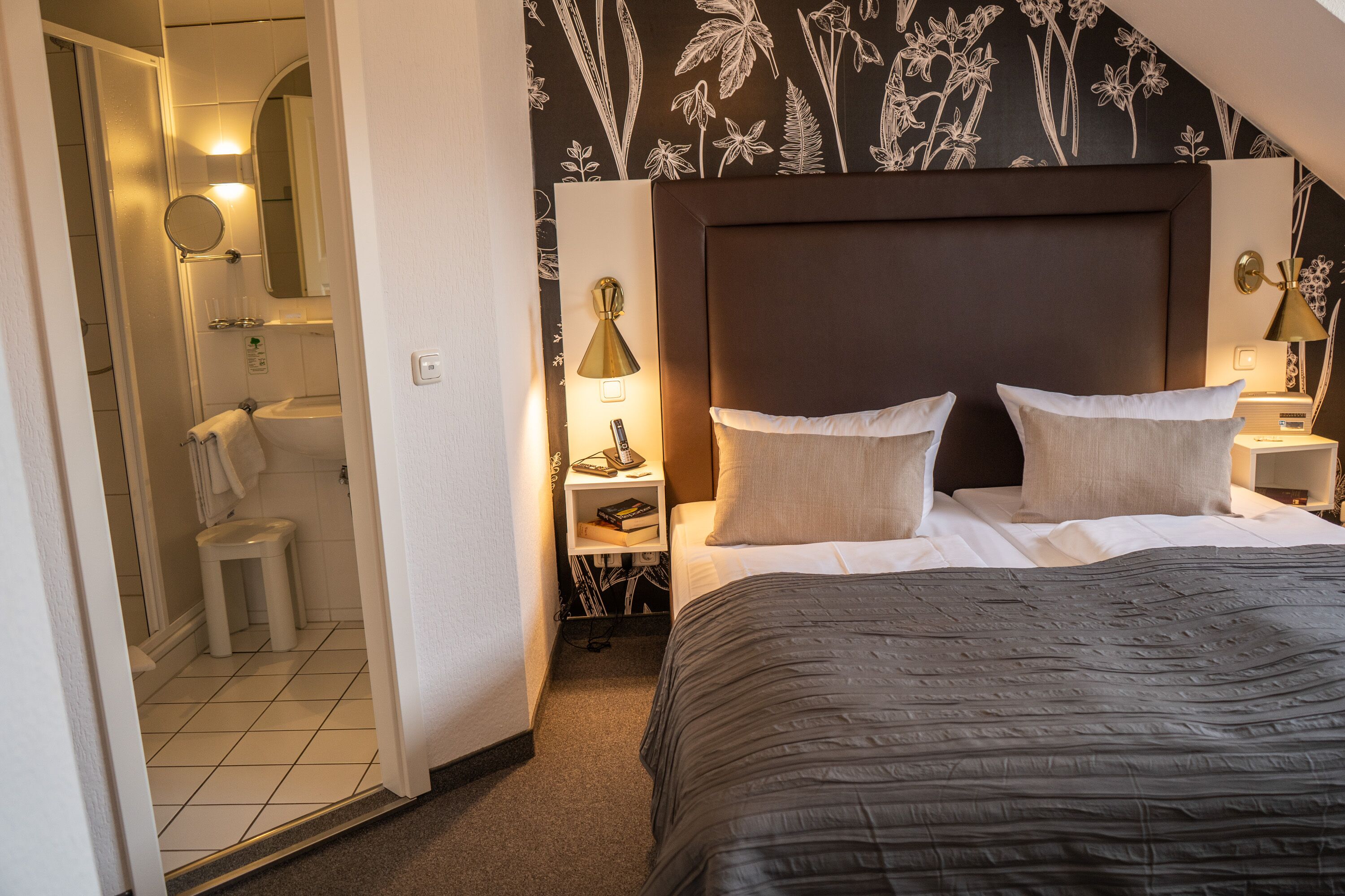 A hotel room with a large bed, a nightstand with a lamp, a bathroom with a toilet and sink, and a shower.