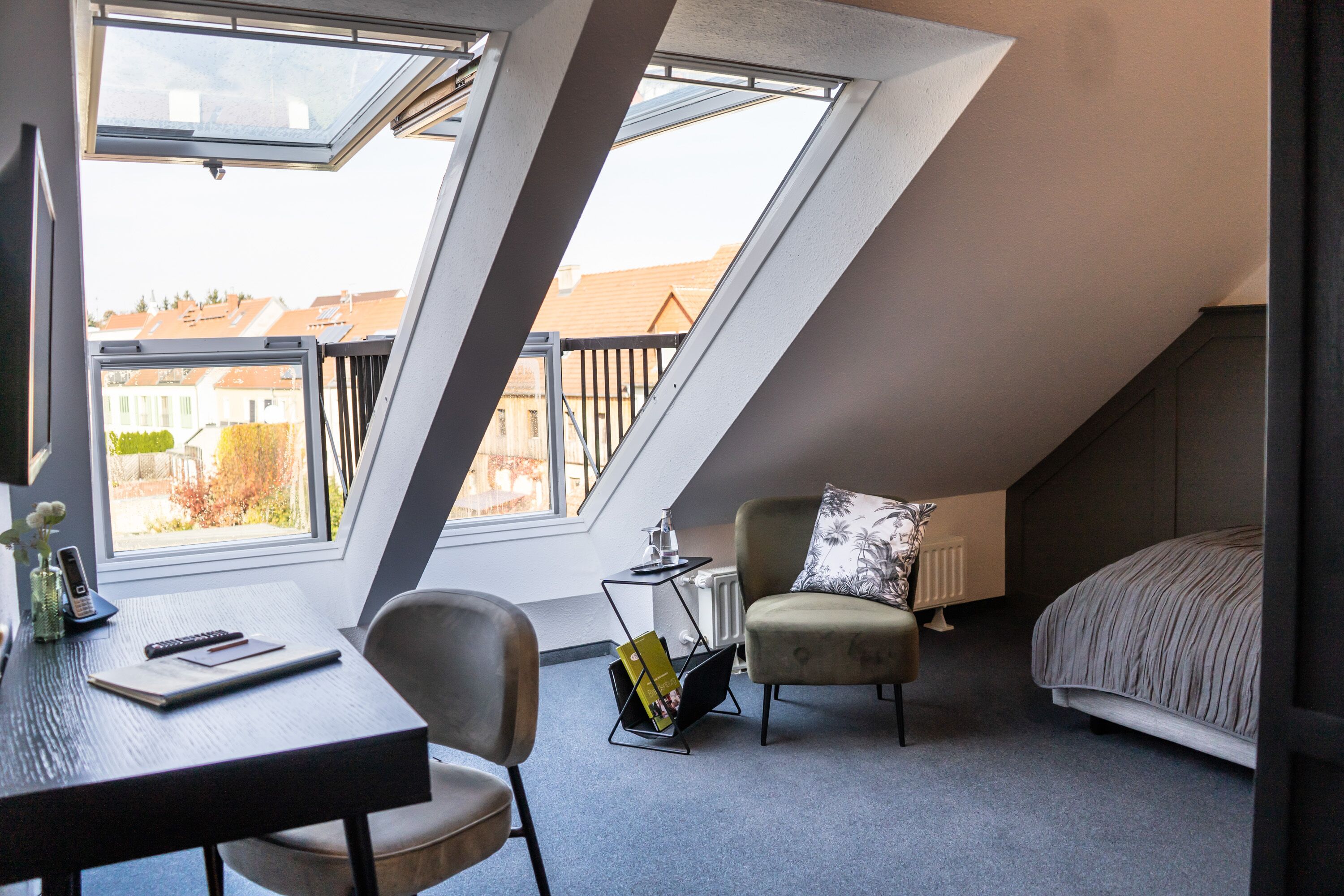 Una habitación con techo inclinado, una cama, un escritorio con silla, una mesita con lámpara, y vistas a edificios a través de amplios ventanales.