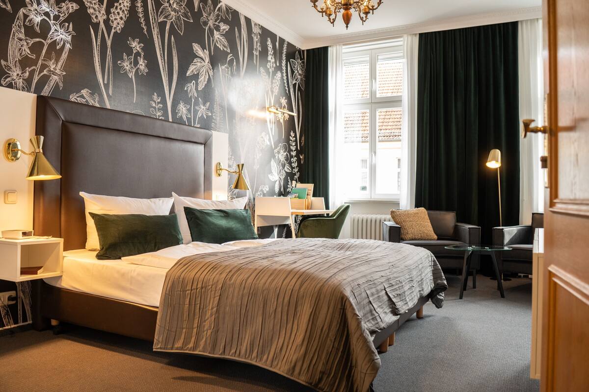 Quarto de hotel com uma cama grande, uma escrivaninha, uma cadeira, um radiador e uma janela com cortinas.