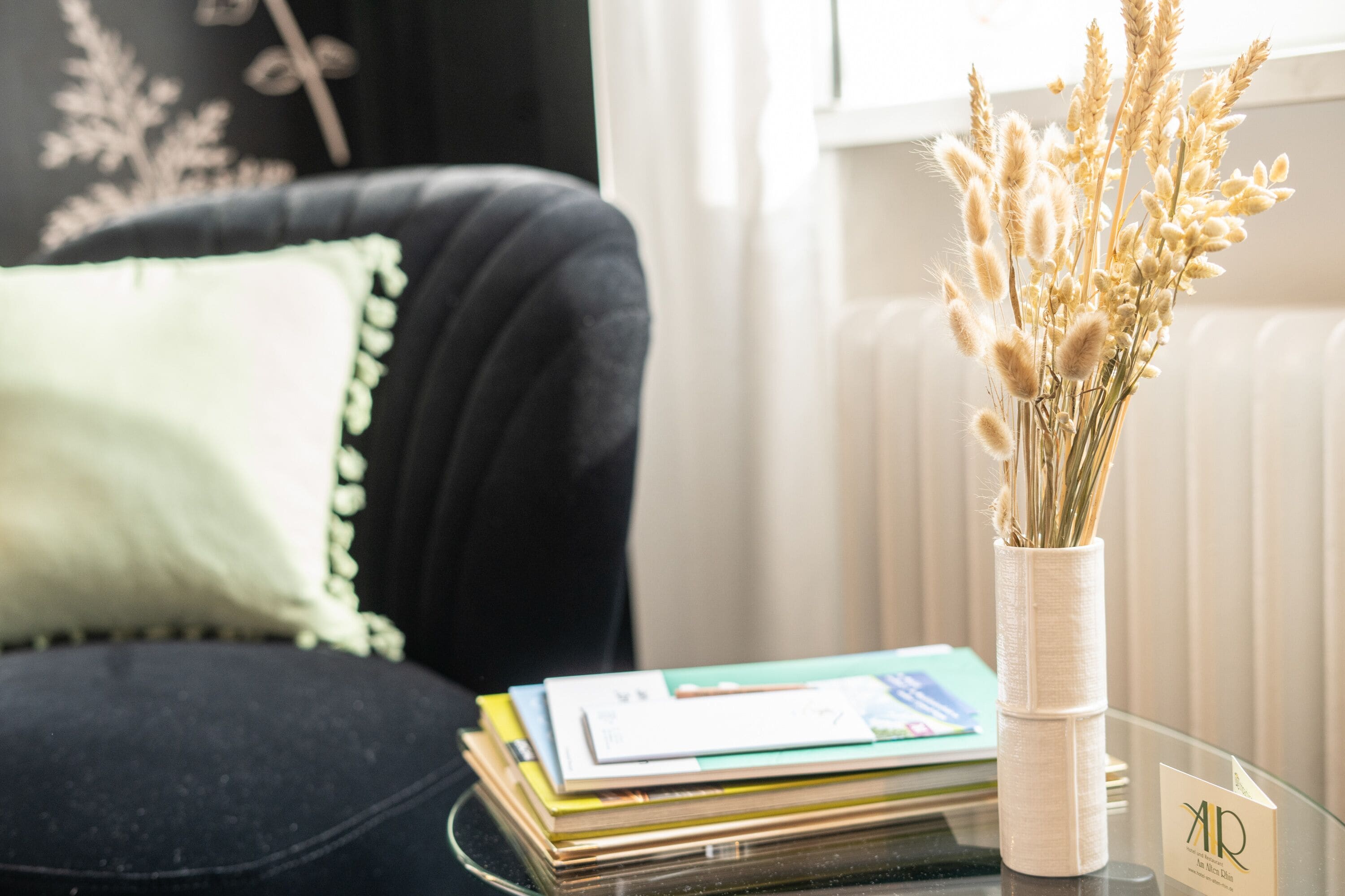 Uma cadeira preta com almofada verde, uma mesa de vidro com um vaso de flores secas e uma pilha de livros.