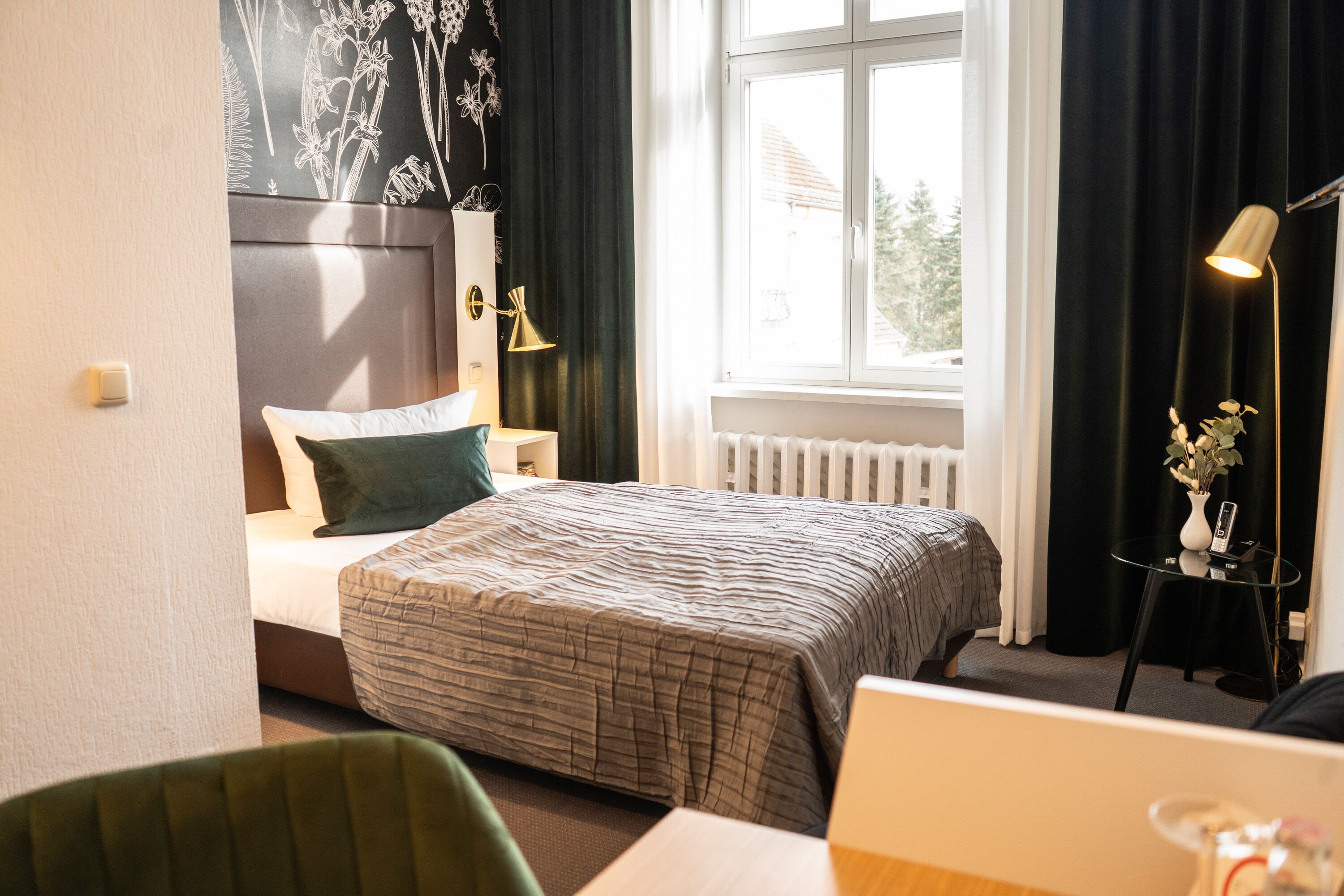 A hotel room with a bed, a desk, a chair, a lamp, and a window with curtains.
