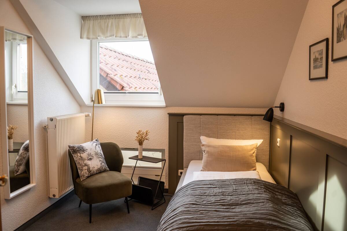 Um quarto com uma cama, uma cadeira, uma mesinha com um vaso e uma janela com vista para um edifício.