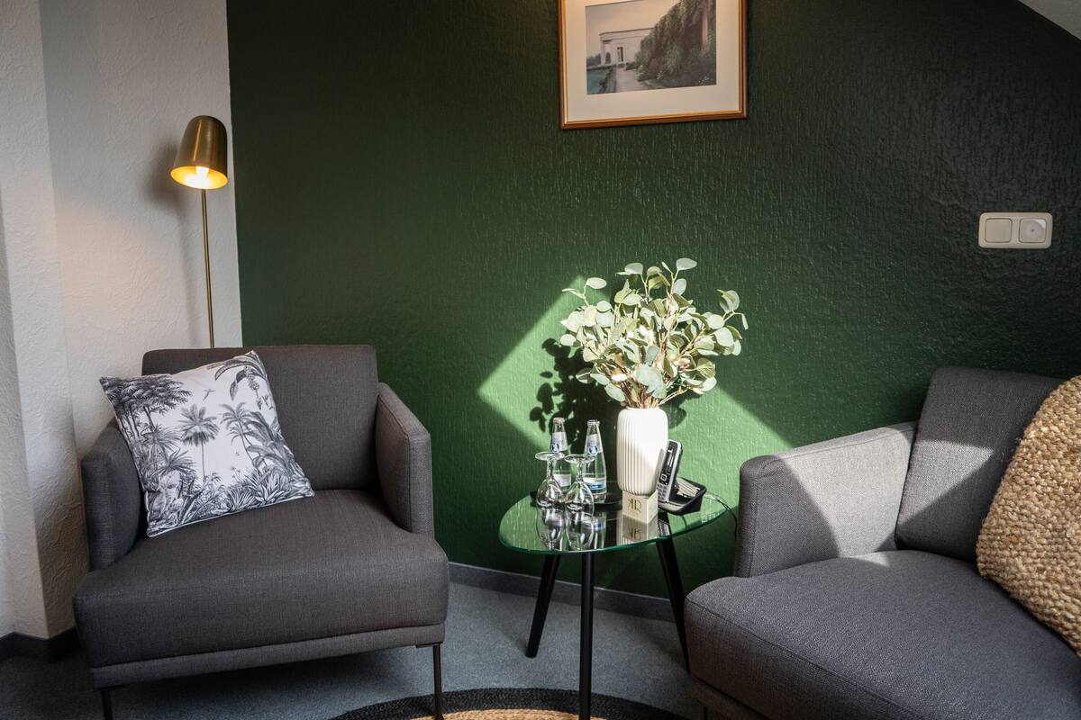 Uma sala de estar moderna com dois sofás cinzas, uma mesa de centro de vidro, um vaso branco com plantas e um quadro na parede.