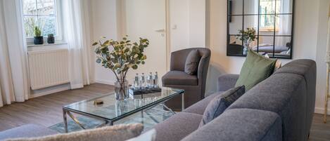 Una sala de estar moderna con un sofá gris, una mesa de centro de cristal y una silla. Se ve una ventana con cortinas y un radiador.