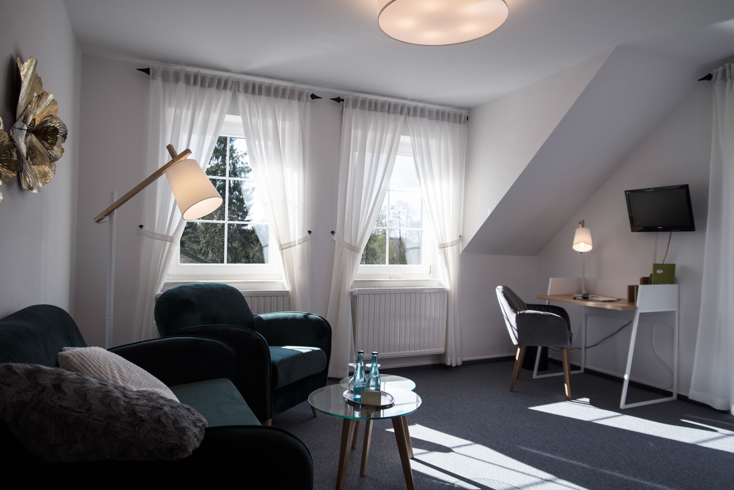 Un salon moderne avec un canapé, une table basse en verre et un bureau avec une chaise. Il y a deux fenêtres avec des rideaux légers, un téléviseur à écran plat fixé au mur et un élément décoratif mural.