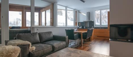 A modern living room with a leather sofa, a wooden coffee table, and a dining area with a table and chairs. Large windows provide natural light and views of the outdoors.