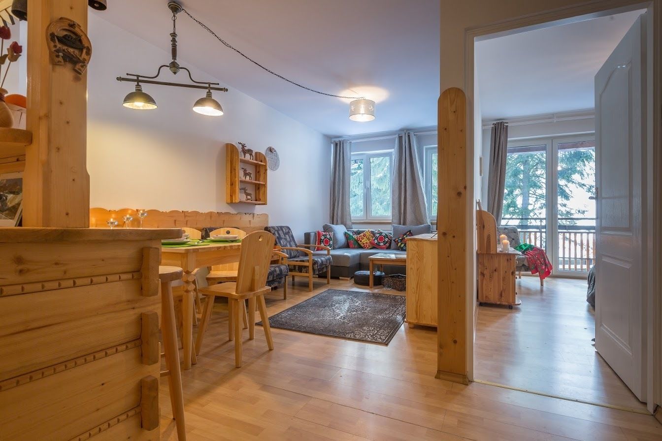 A cozy living room with a wooden dining table, chairs, a sofa, and a coffee table. There are two windows with curtains, a wooden bookshelf, and a hanging light fixture.