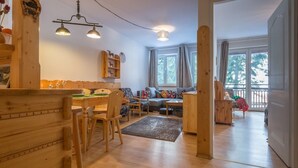 Tunnelmallinen olohuone, jossa on puinen ruokapöytä, tuoleja, sohva ja sohvapöytä. Huoneessa on kaksi ikkunaa, joissa on verhot, puinen kirjahylly ja riippuvalaisin.