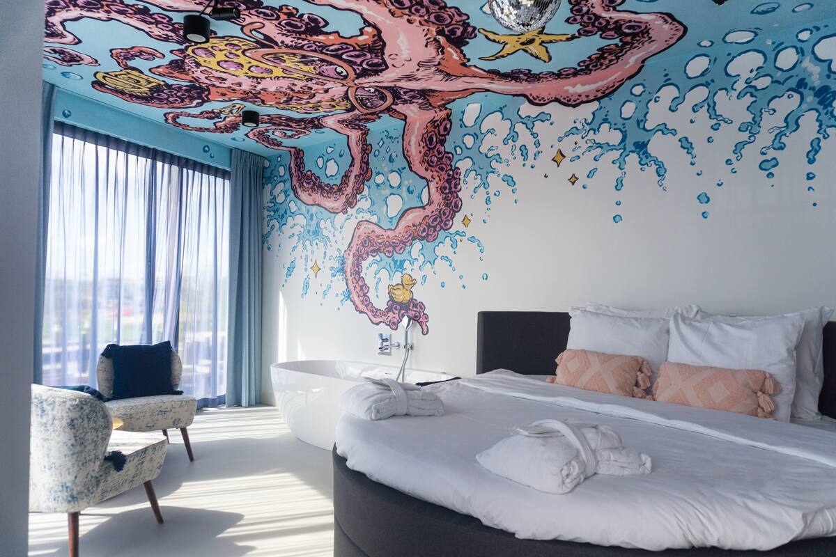 Moderni hotellihuone, jossa on suuri sänky, oleskelualue ja kattoon maalattu koristekuva.