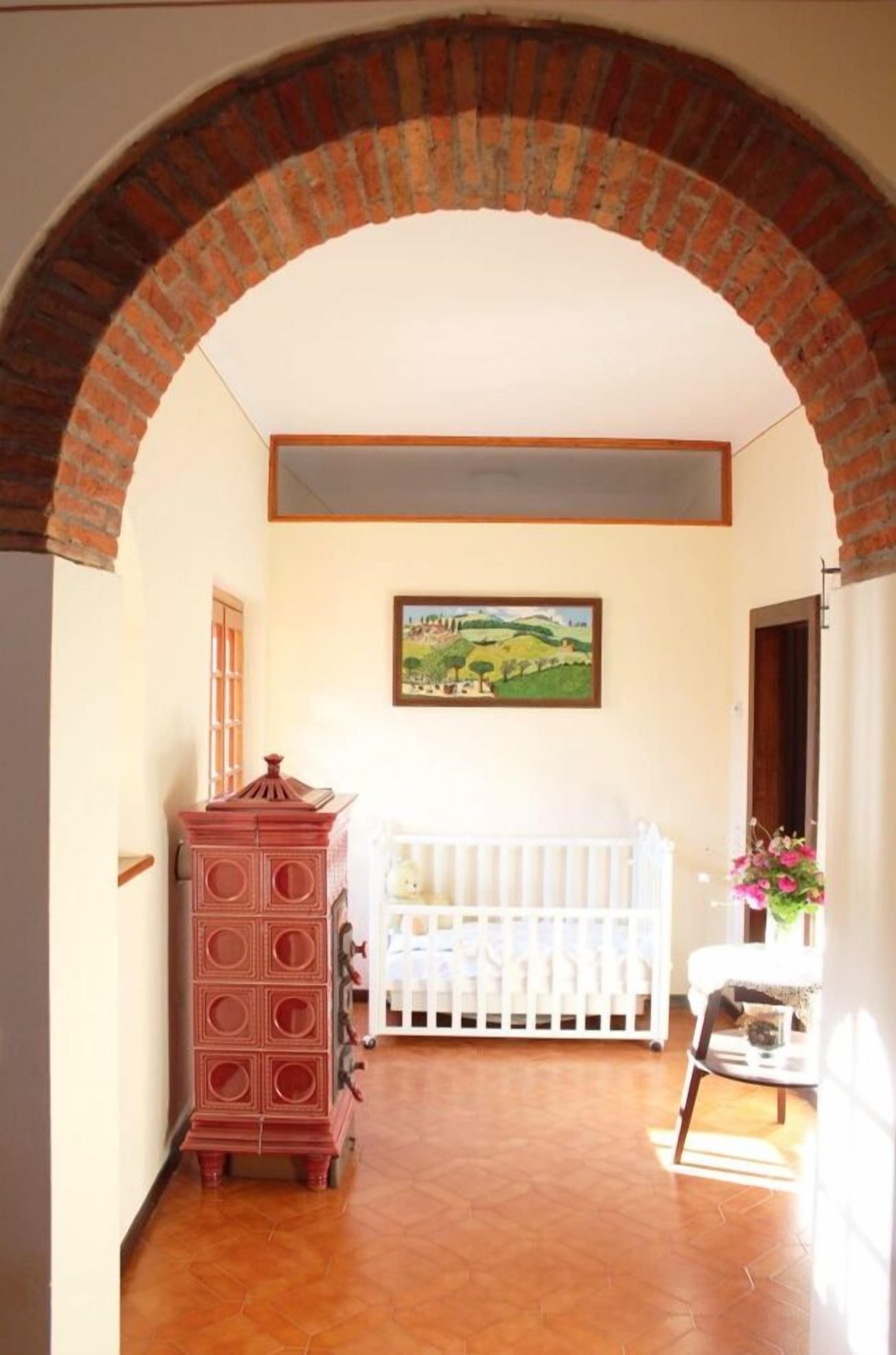 A hallway with a brick archway, a white crib, a red cabinet, and a painting on the wall.