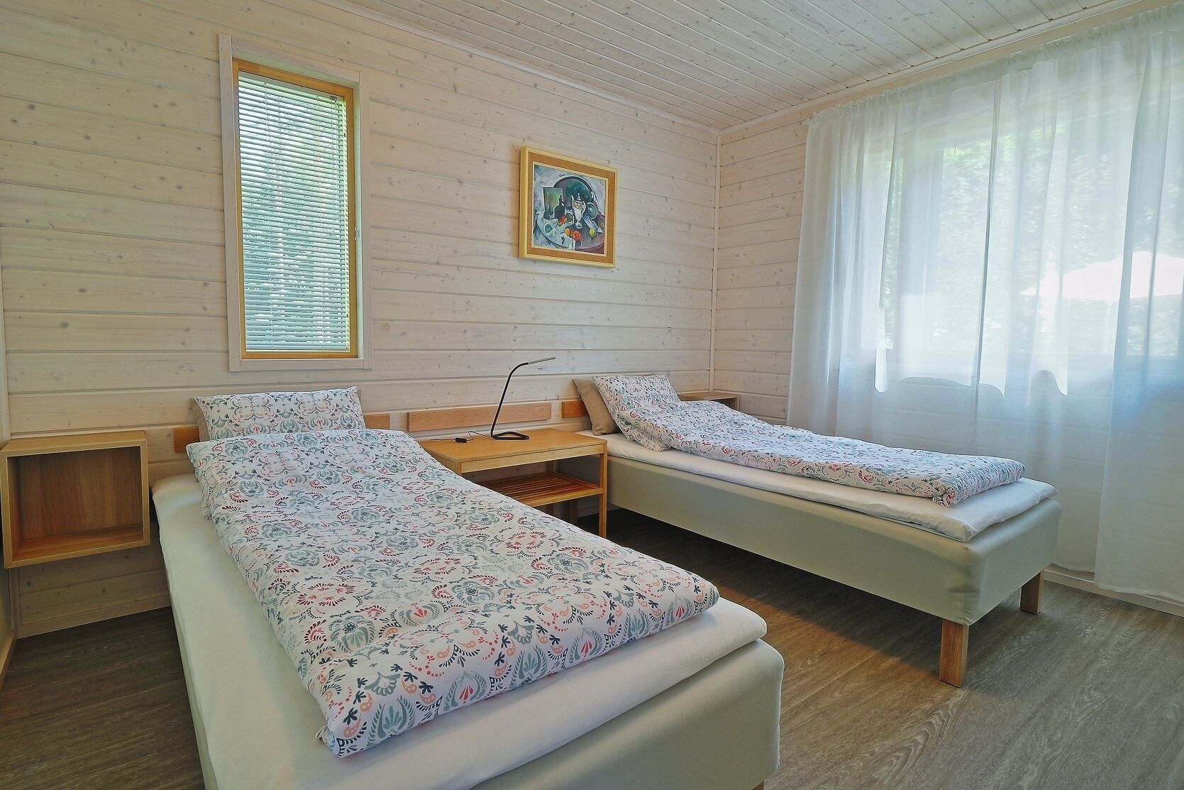 A room with two single beds, wooden walls, a small table, a nightstand, a picture on the wall, and a window with curtains.