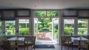 Un comedor con mesas y sillas, ventanas amplias con vistas a un jardín y una puerta que da al exterior.