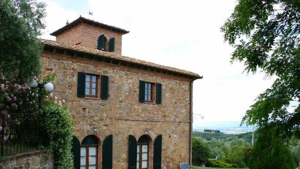 A stone building with a tiled roof, multiple windows with shutters, and a small garden area with a bench and potted plants.