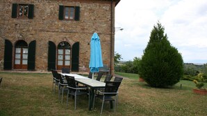 A stone building with a dining area featuring a table and chairs, a blue umbrella, and a large tree in the background.