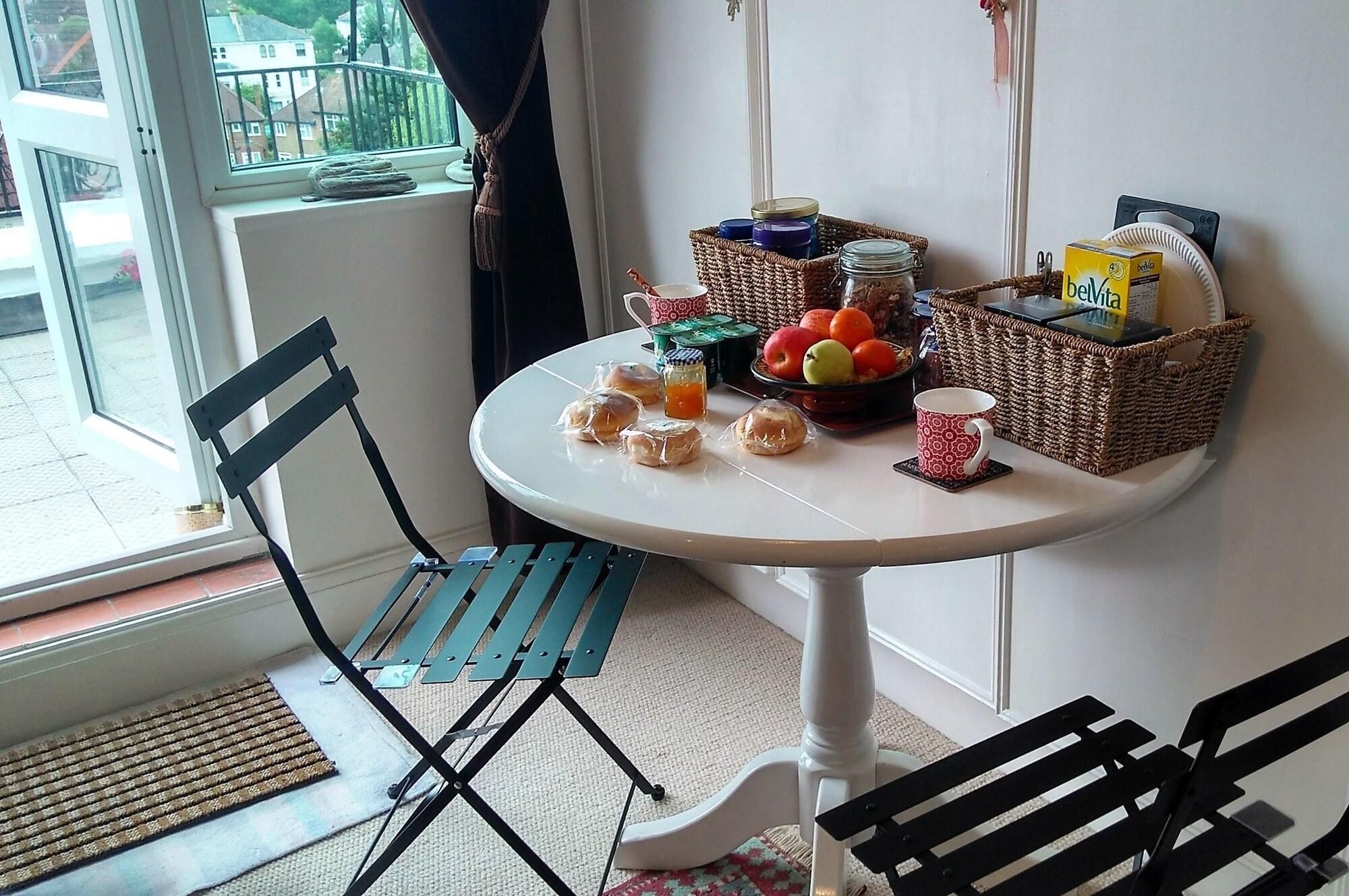 A corner room with a table set with bread, apples, and a cup, a wicker basket, and a window with curtains.