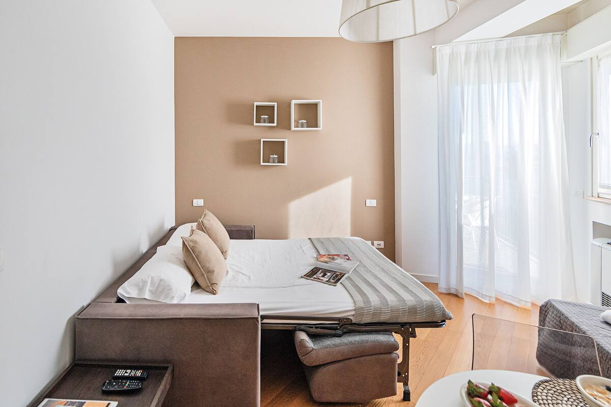 Moderni hotellihuone, jossa on sänky, pieni pöytä, tuoli ja ikkuna, jossa on verhot.