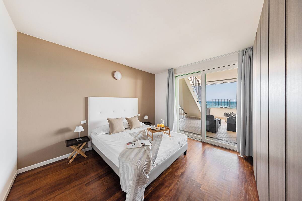 Moderni hotellihuone, jossa on suuri sänky, yöpöytä lampun kanssa, tarjotinalus, jossa on appelsiinimehua, ja liukuoven kautta avautuva näkymä rannalle.