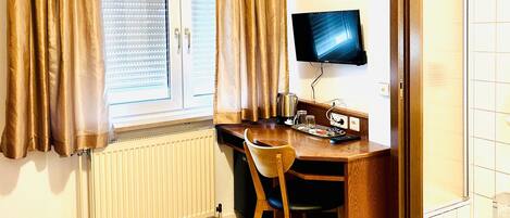 Une pièce avec un bureau en bois, une chaise, un téléviseur fixé au mur et une fenêtre avec des rideaux.