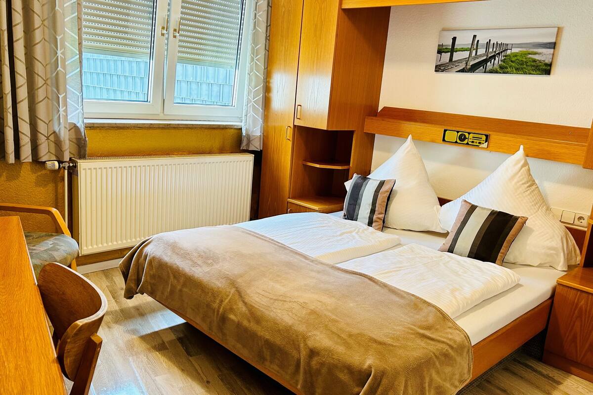 A hotel room with a bed, a desk, a chair, a radiator, a window with blinds, and a picture on the wall.