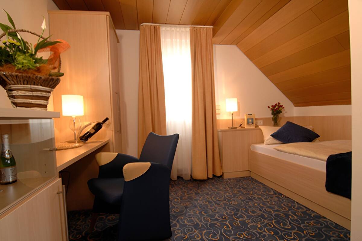 A small, cozy room with a bed, a desk with a lamp, a chair, and a window with curtains.