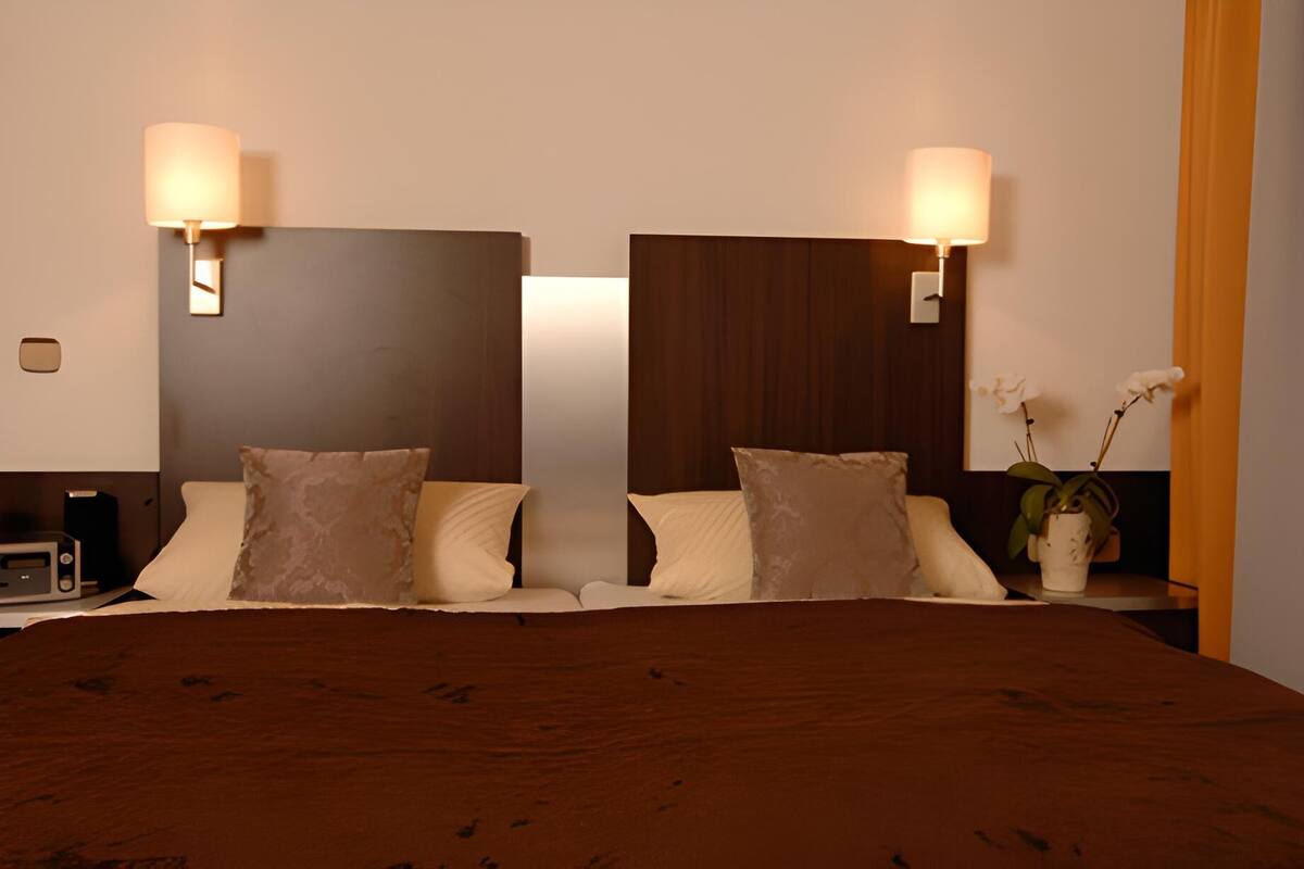 A hotel room with a large bed, two wall-mounted lamps, a nightstand with a plant, and a wooden headboard.