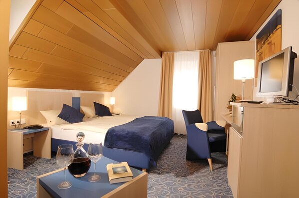 A hotel room with a wooden ceiling, a bed with blue bedding, a desk with a lamp, a TV, and a minibar. - Garni-Hotel Adler (Ostfildern)