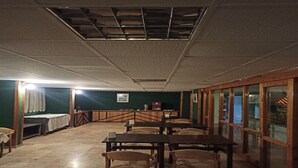 Una habitación con techo alto, ventanas de madera y suelo de baldosas. Varias mesas y sillas dispuestas para comer.