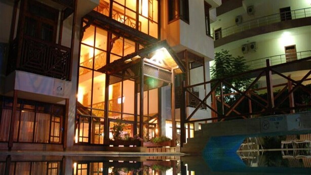 Un edificio de varios pisos con amplios ventanales y un balcón, que cuenta con una zona de piscina, sillas y plantas.