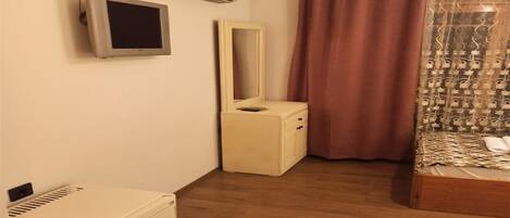 Quarto compacto com piso de madeira, mesa de cabeceira pequena, cama com roupa de cama estampada, espelho e ar-condicionado fixado na parede.
