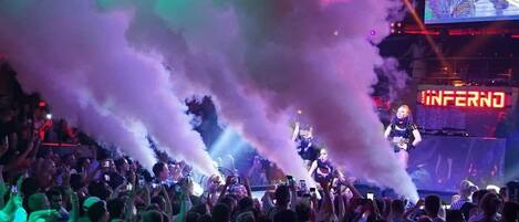 A crowded indoor event with a stage, smoke effects, and a large screen displaying "INFERNO".