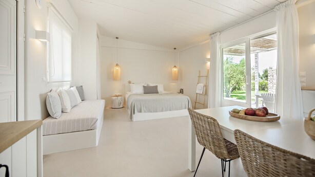 A bright, white room with a bed, a dining table with fruit, and a view of greenery outside. - Ta Spitakia (Kos)