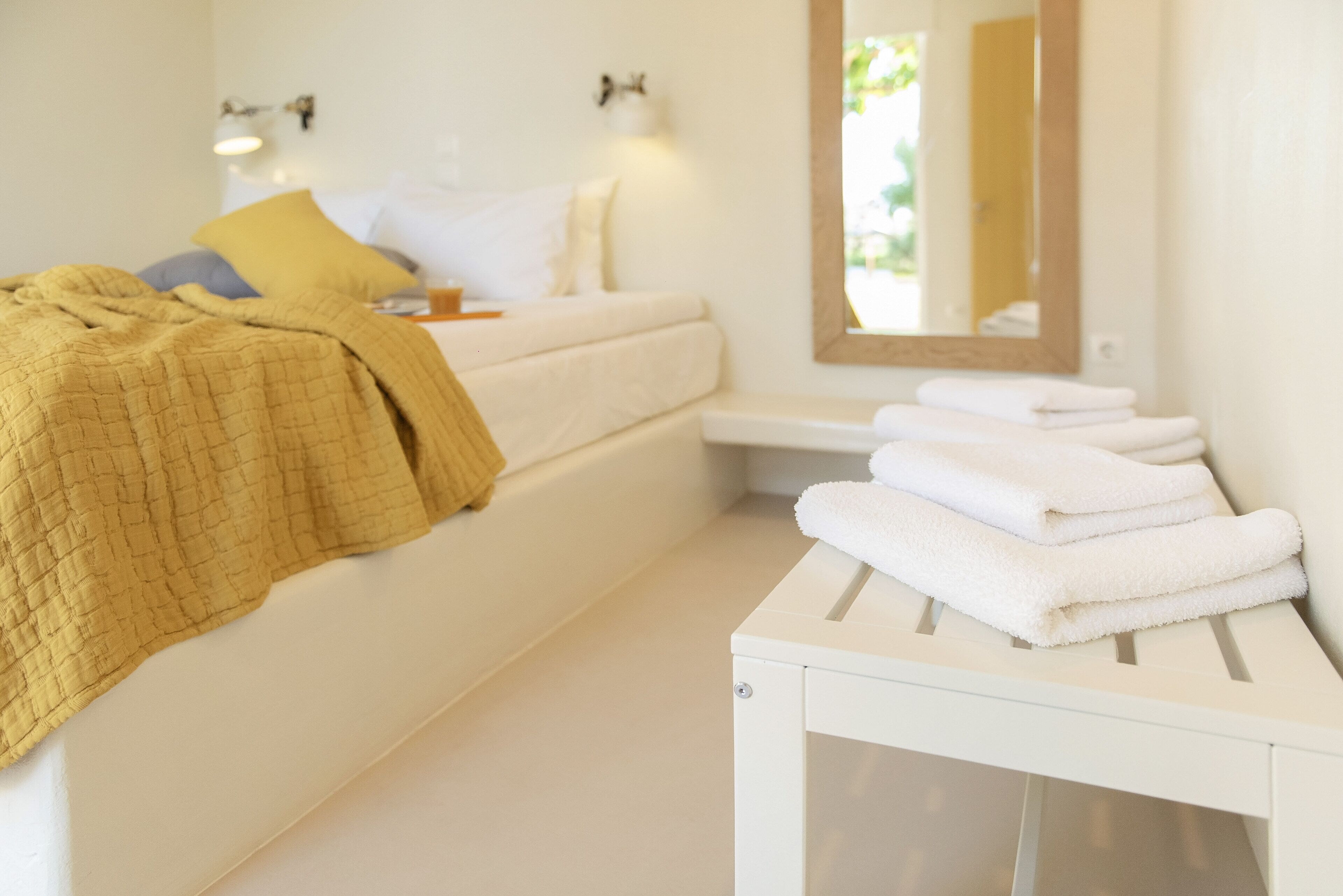 A hotel room with a bed, a bedside table with towels, a mirror, and a wall-mounted light fixture.