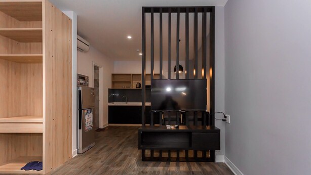 A modern living room with a built-in TV, wooden shelving, and a kitchen in the background. - VN HOUSE Beach (Da Nang)