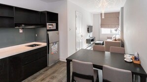 A modern kitchen with dark cabinetry, a built-in oven, and a refrigerator. There is a dining area with a table and chairs, and a living space with a sofa and a TV. - VN HOUSE Beach (Da Nang)