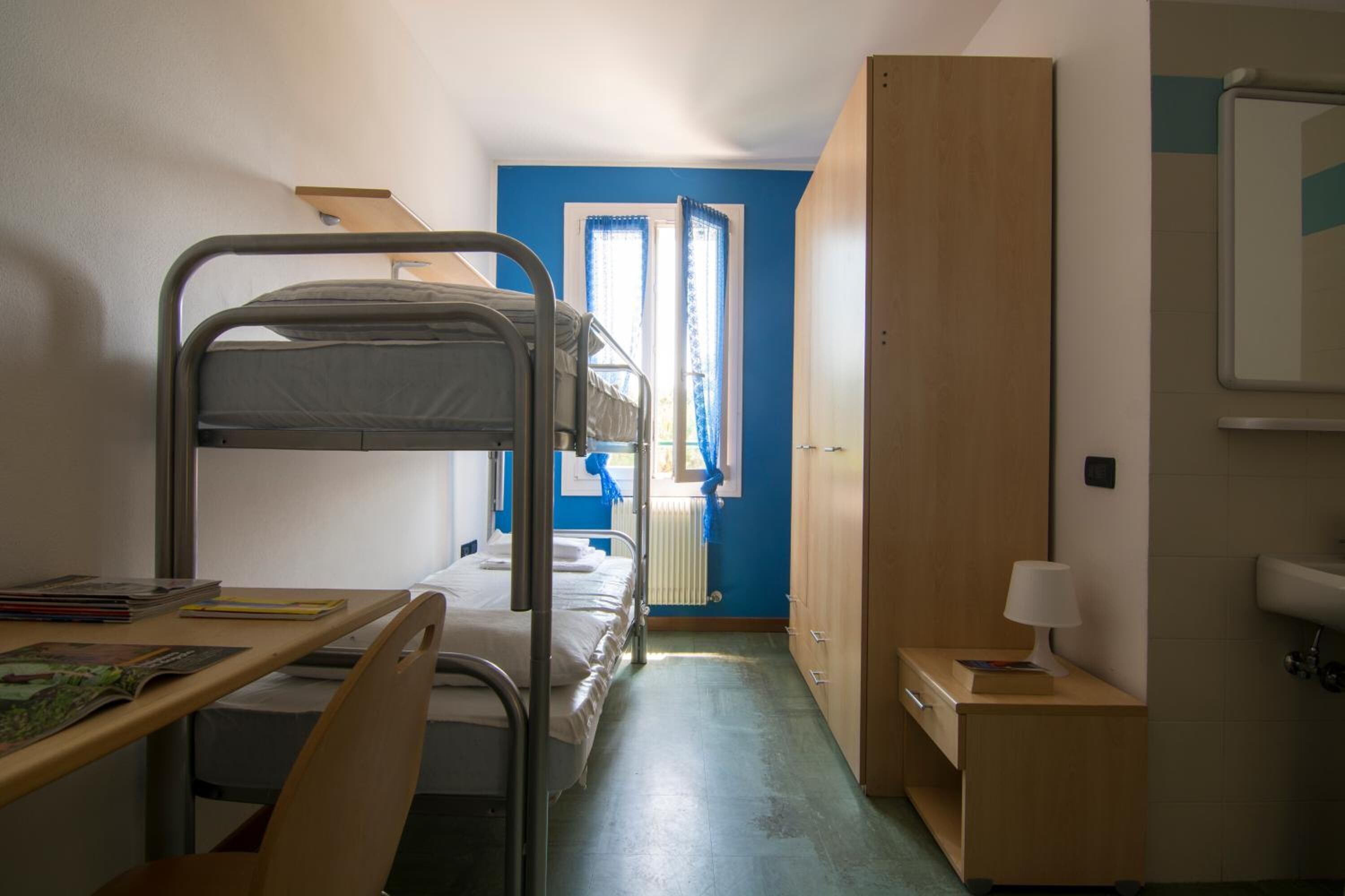 A dormitory room with bunk beds, a desk, a chair, a small cabinet, and a window with curtains.