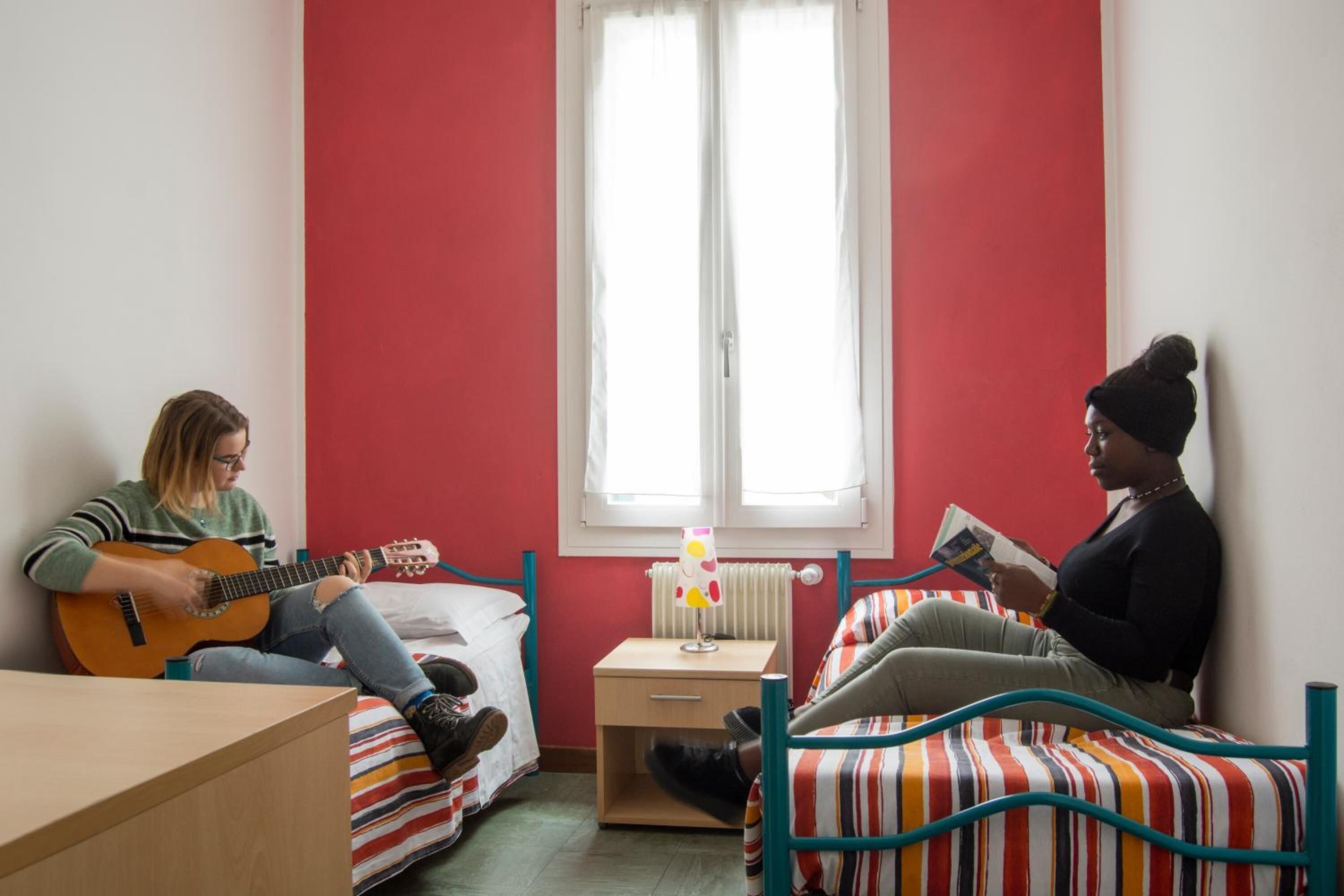 A room with two beds, one with a striped bedspread and another with a plain one. There is a window with white curtains, a bedside table with a lamp, and a guitar on one bed.