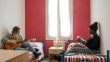 A room with two beds, one with a striped bedspread and another with a plain one. There is a window with white curtains, a bedside table with a lamp, and a guitar on one bed.