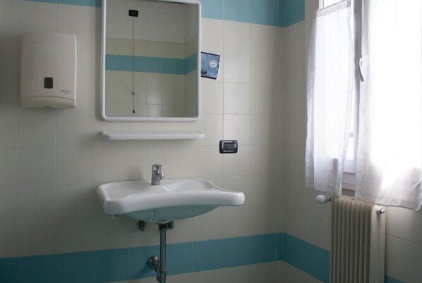 A bathroom with a white sink, a mirror, a soap dispenser, and a window with a white curtain. - Ostello Bassano Del Grappa (Bassano del Grappa)