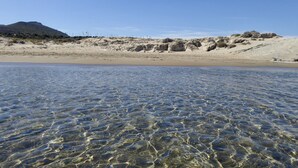 A beach with clear shallow waters, sandy shore, and rocky hills in the background.