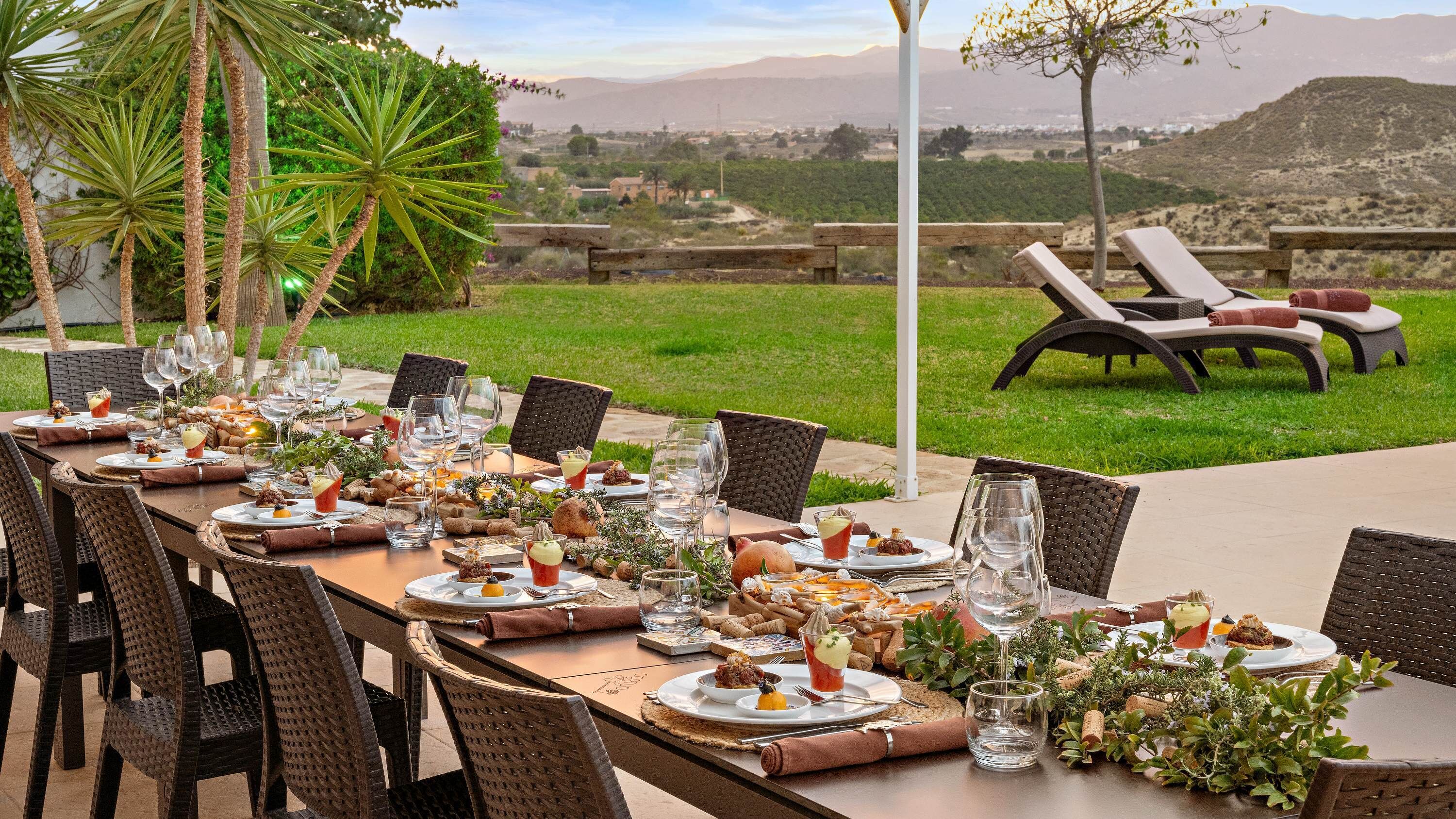 Un espacio de comedor al aire libre con una larga mesa dispuesta para una comida, rodeada de sillas y con vistas a un paisaje distante.