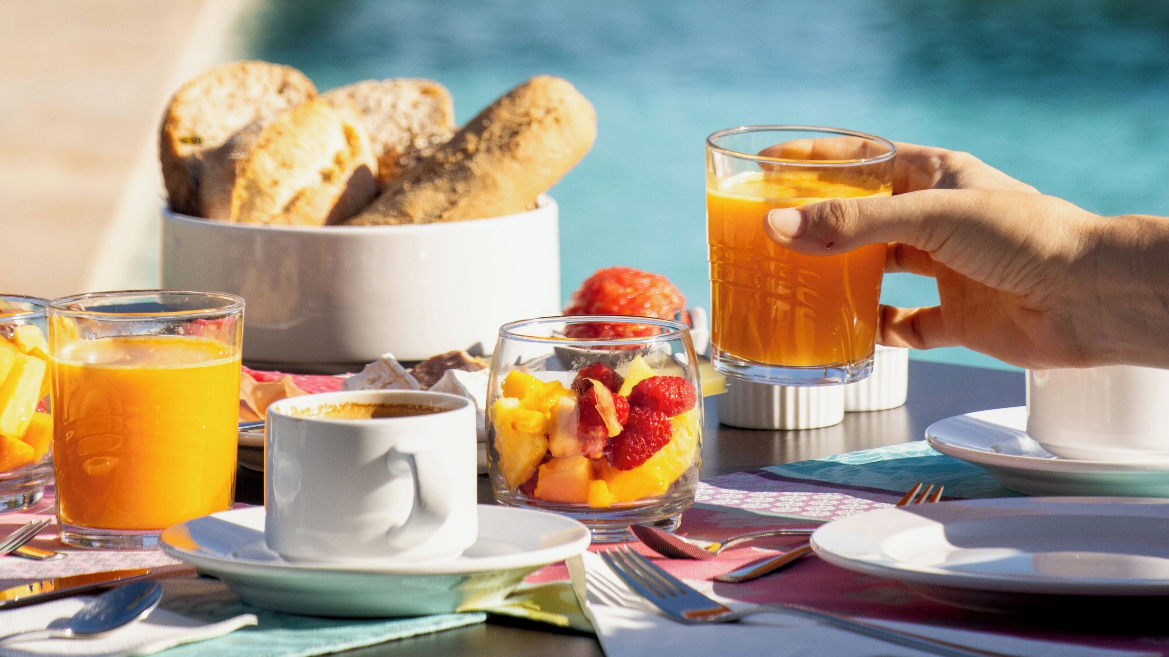 A breakfast setting with fruit, orange juice, and pastries by a pool.