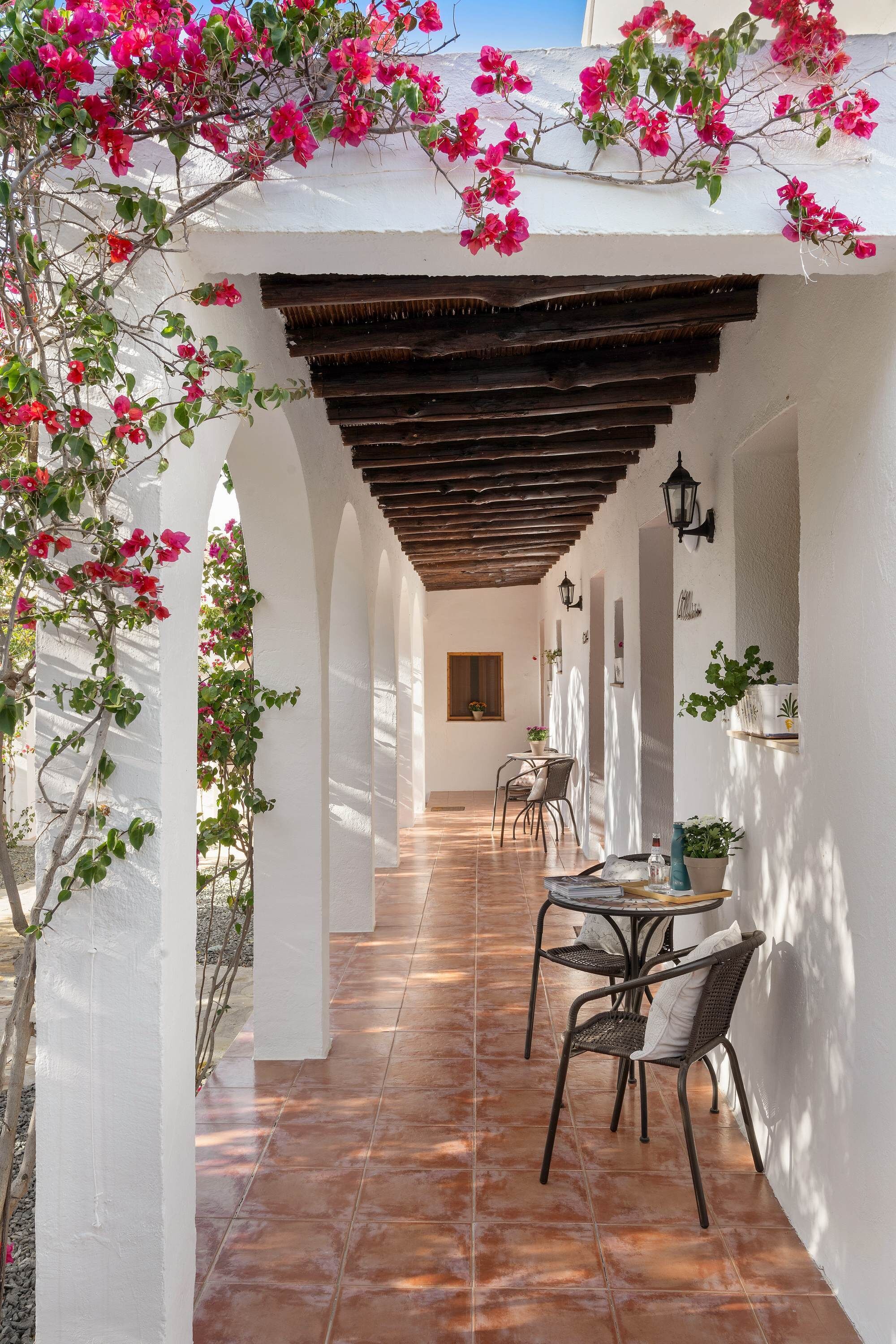 A covered outdoor area with a tiled floor, white walls, wooden beams, and potted plants.